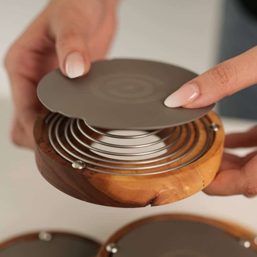 دم افزار اسپرو (Sprew) تصویری نزدیک از دستها که در حال قرار دادن درپوش خاکستری بر روی دم افزار اسپرو هستند. فنر دستگاه در پایه چوبی آن جمع شده و درپوش در حال بسته شدن است.