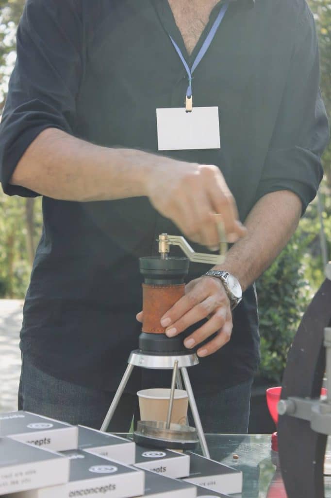 ایستگاه یکم: تلسی فردی در یک رویداد در فضای باز، در حال آسیاب کردن قهوه با آسیاب دستی و ریختن آن مستقیماً در پرتافیلتر یک دستگاه اسپرسوساز دستی است. در پیشزمینه، جعبههای محصول با لوگوی onconcepts دیده میشود.