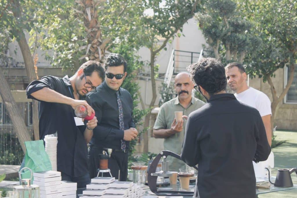 ایستگاه یکم: تلسی گروهی از مردان در یک رویداد در فضای باز، دور یک میز جمع شدهاند و در حال تماشای نمایش زنده دمآوری قهوه هستند. روی میز، محصولات و جعبههای برند onconcepts قرار دارد.