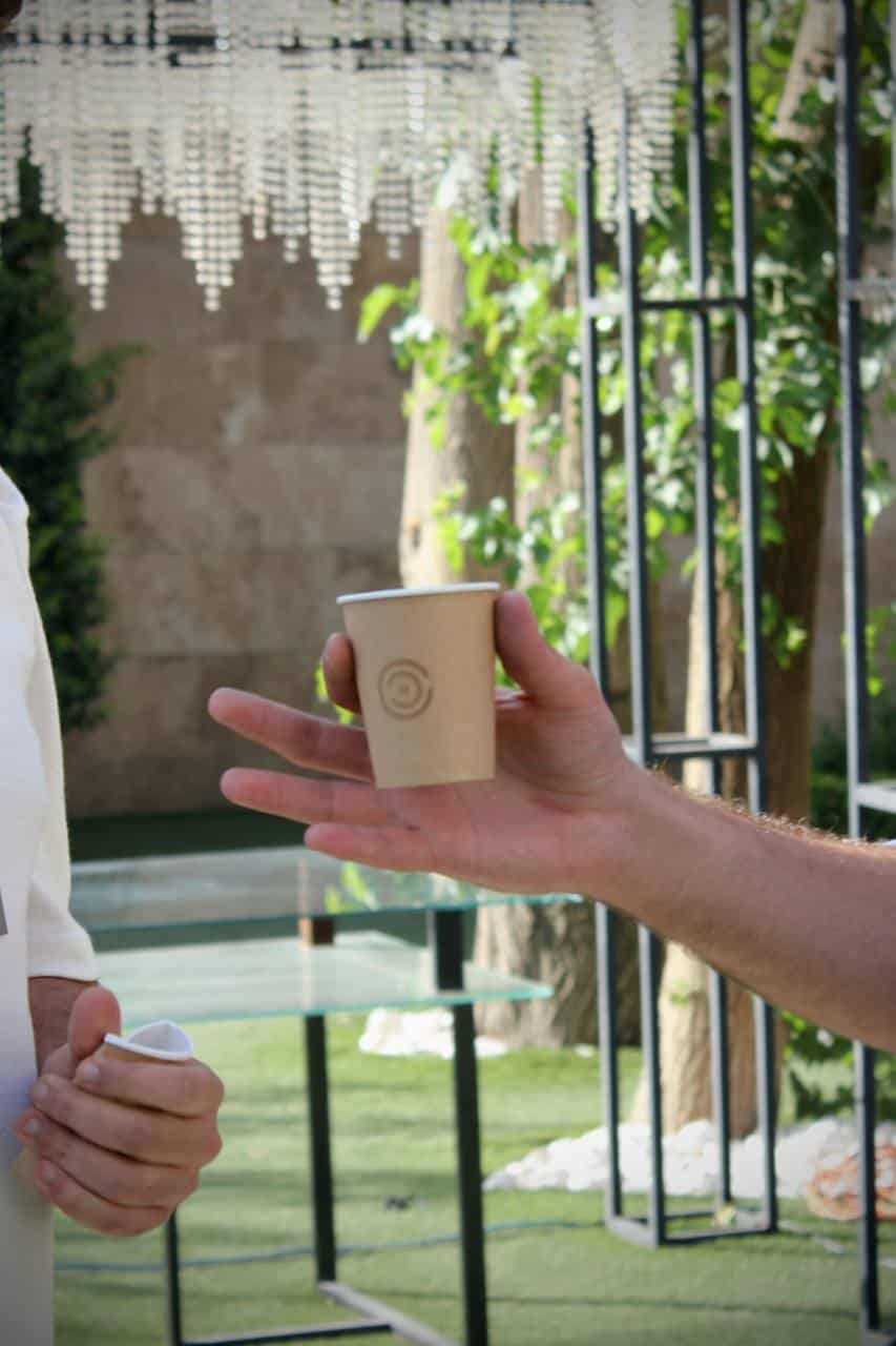 ایستگاه یکم: تلسی تصویری نزدیک از یک دست که فنجان قهوه کاغذی با لوگوی onconcepts را به شخص دیگری تعارف میکند. این صحنه در یک رویداد در فضای باز و سرسبز اتفاق میافتد.